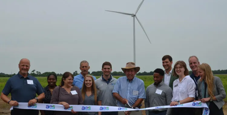 A group of people standing outdoors in front of a wind turbine, smiling and holding a ribbon with the AES logo. One person is cutting the ribbon, marking a ceremonial event. The background shows a grassy field and a cloudy sky.