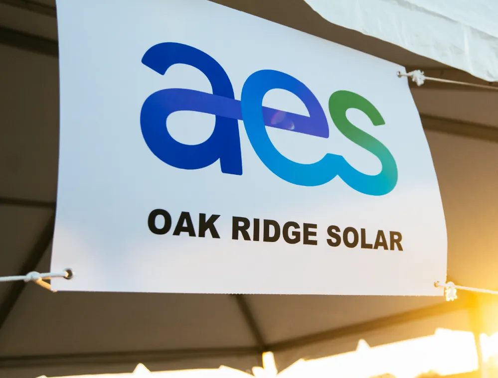 A banner displaying the logo 'aes' in blue and green, with the text 'OAK RIDGE SOLAR' underneath, hanging from a tent with a sunset in the background.