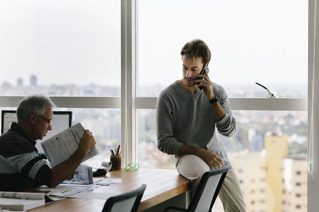 Two men in a windowed office talking; one is sitting on the desk on the phone.