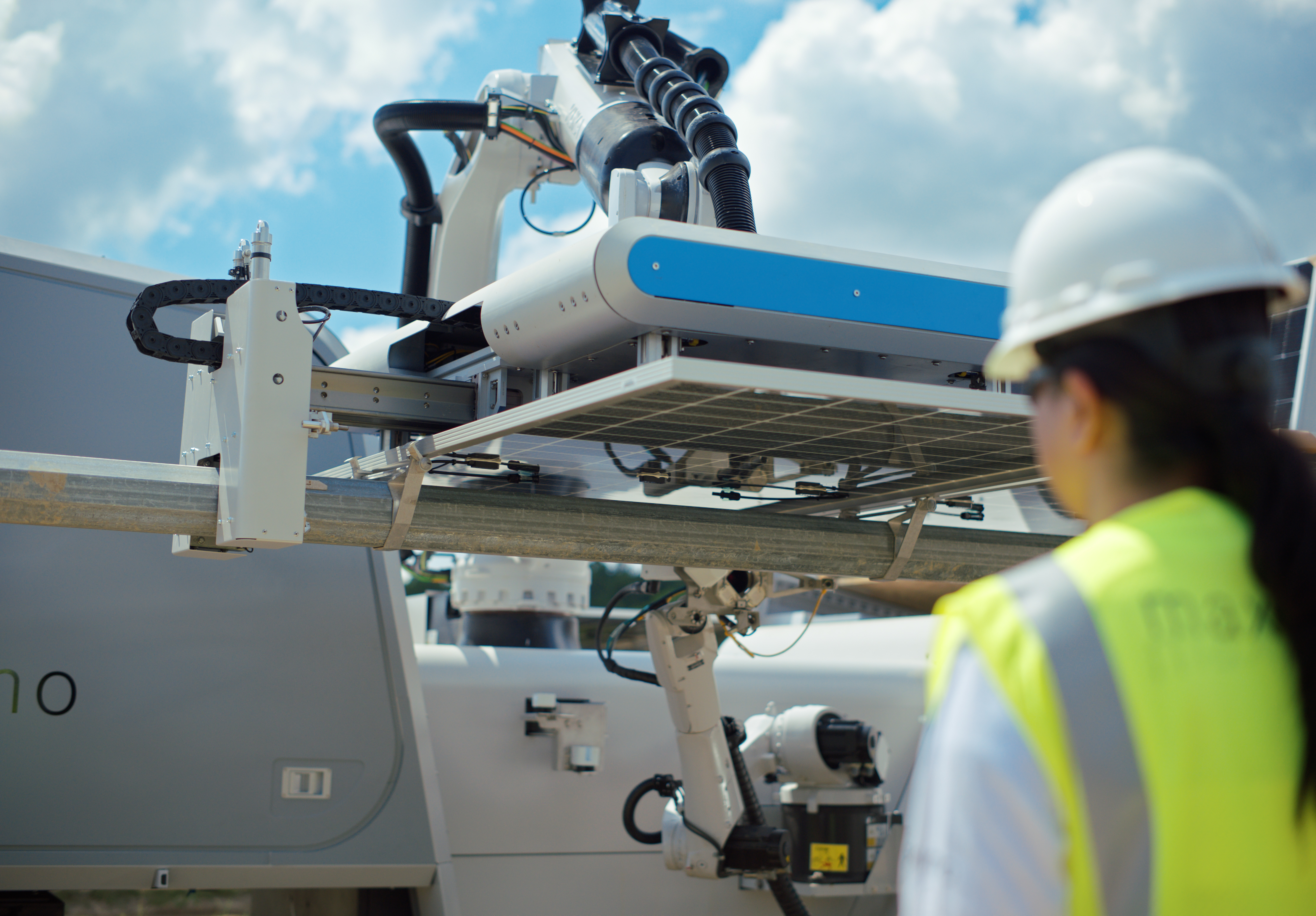 A worker in a hard hat and high-visibility vest observes a robotic arm installing a solar panel on a metal structure under a blue sky with clouds.