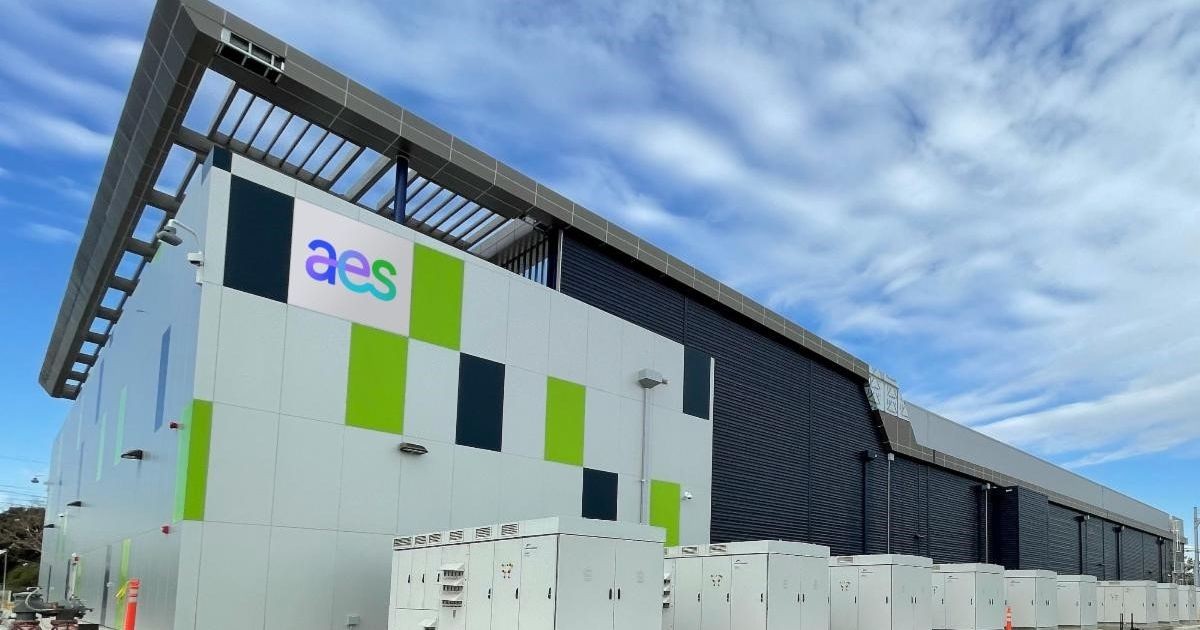 A modern industrial building with a sign displaying 'aes' on its facade. The building features a geometric design with green and dark blue panels. Several white utility boxes are positioned in front. The sky is partly cloudy.