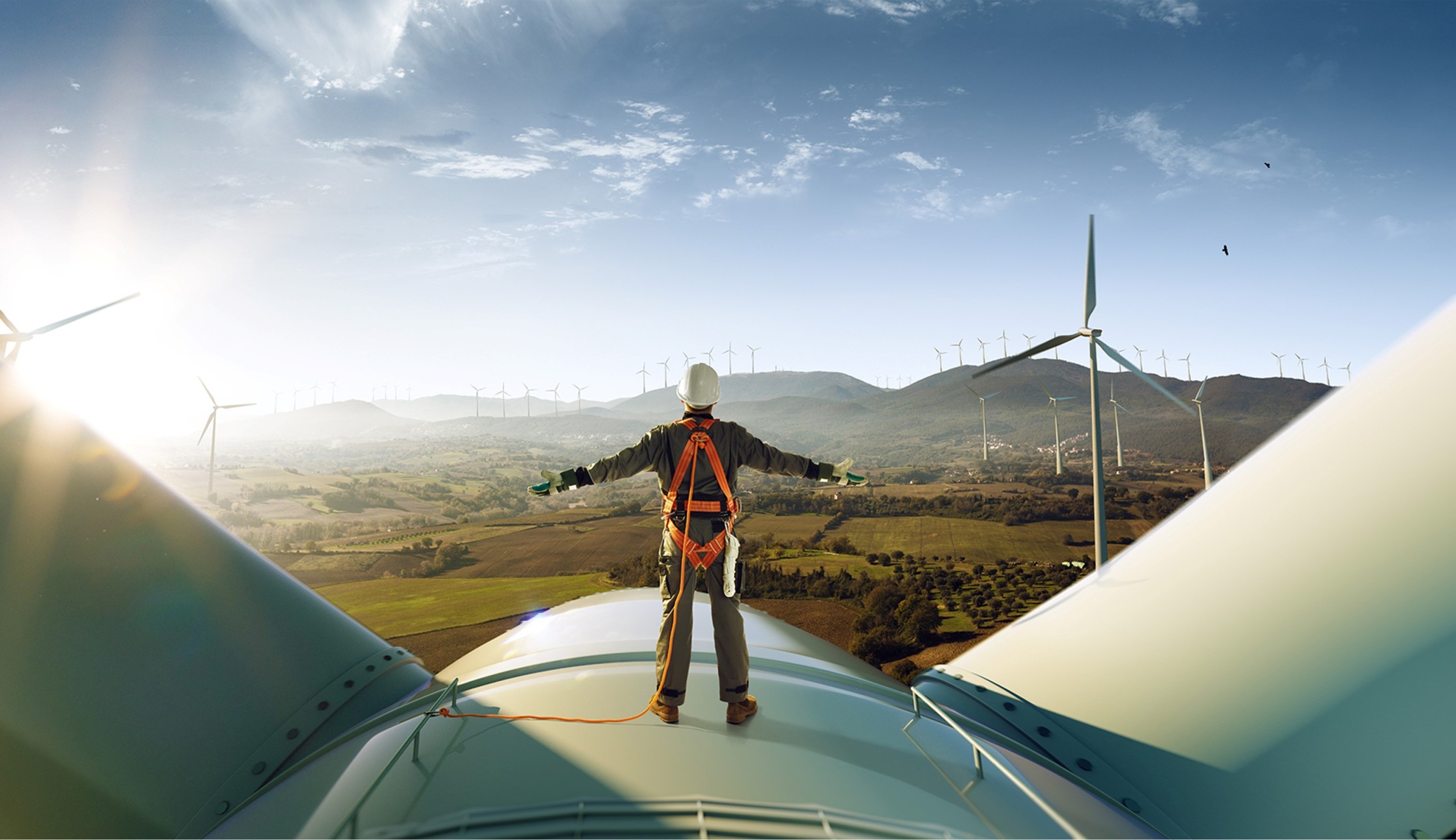 A person wearing a safety harness and helmet stands on top of a wind turbine, overlooking a landscape filled with multiple wind turbines and rolling hills under a blue sky. The sun is setting on the left, casting a warm glow over the scene.