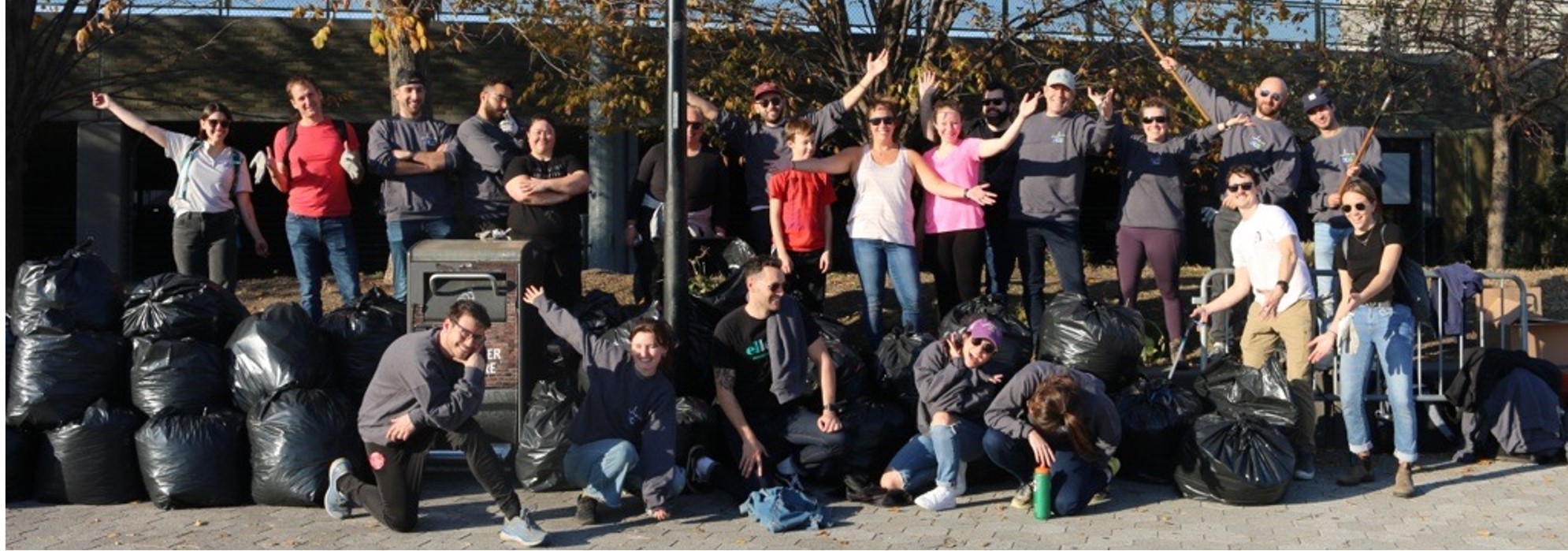 A group of people posing outdoors with black garbage bags piled in front of them. They are smiling and some have their arms raised in celebration. Trees with autumn leaves are in the background.