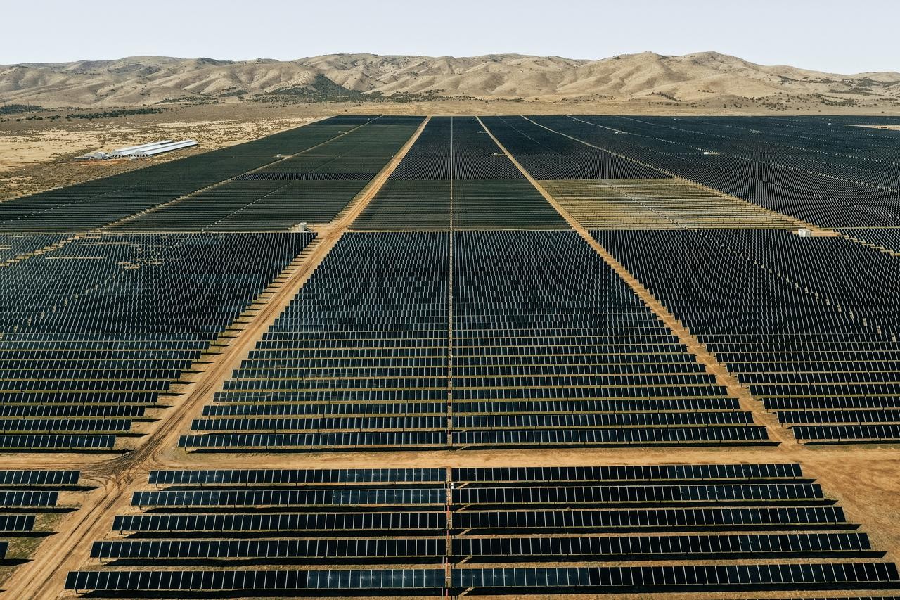 Aerial view of a vast solar farm with rows of solar panels stretching into the distance. The panels are arranged in a grid pattern on a flat, arid landscape. In the background, there are low, barren hills under a clear sky.