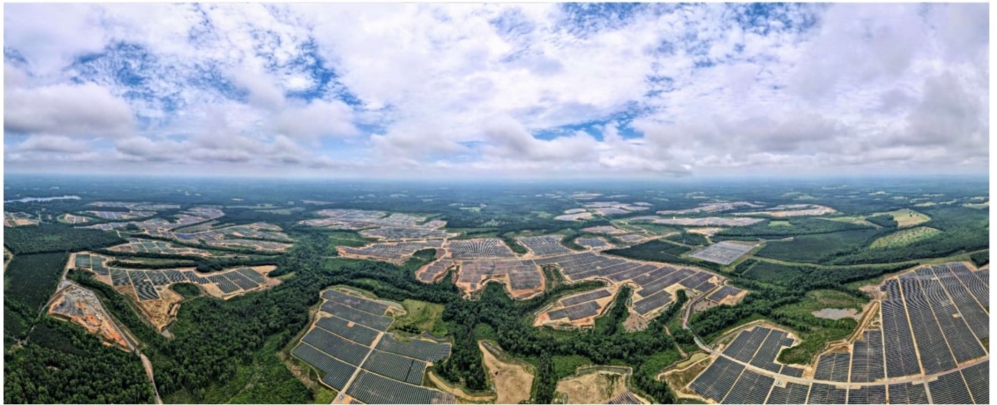 Aerial view of a vast solar farm with numerous solar panels arranged in large sections across a landscape of greenery and forests under a partly cloudy sky.