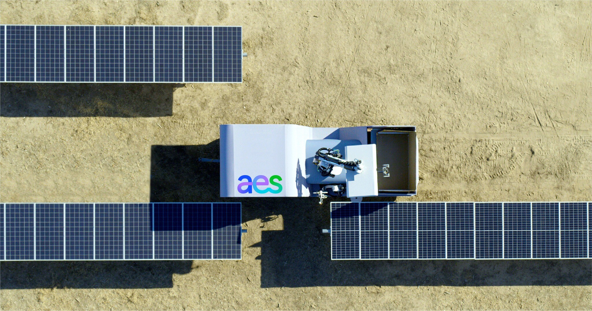 Aerial view of a solar panel installation on a sandy terrain. A robotic device with the AES logo is positioned between two rows of solar panels, performing maintenance or cleaning tasks.