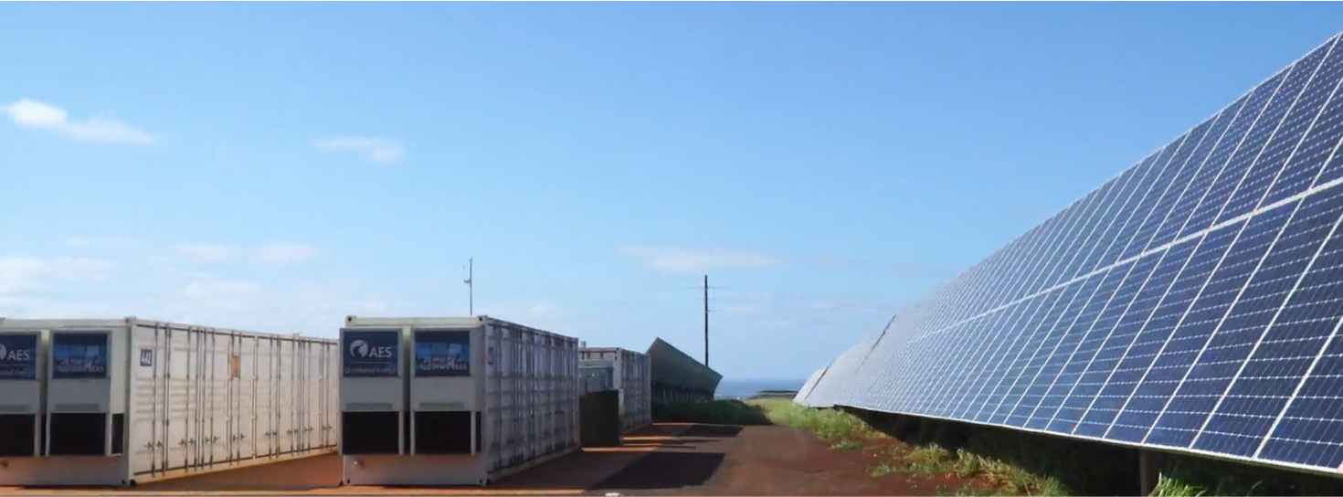 A row of large white containers with AES logos on the left, and a field of solar panels on the right under a clear blue sky.