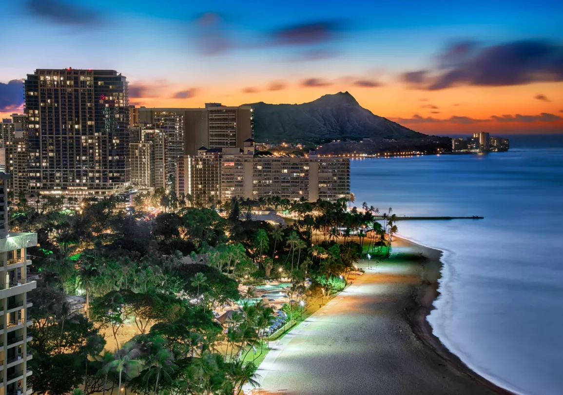 A cityscape at dusk with illuminated buildings near a beach. The ocean is calm, and a mountain is visible in the background under a vibrant sunset sky.