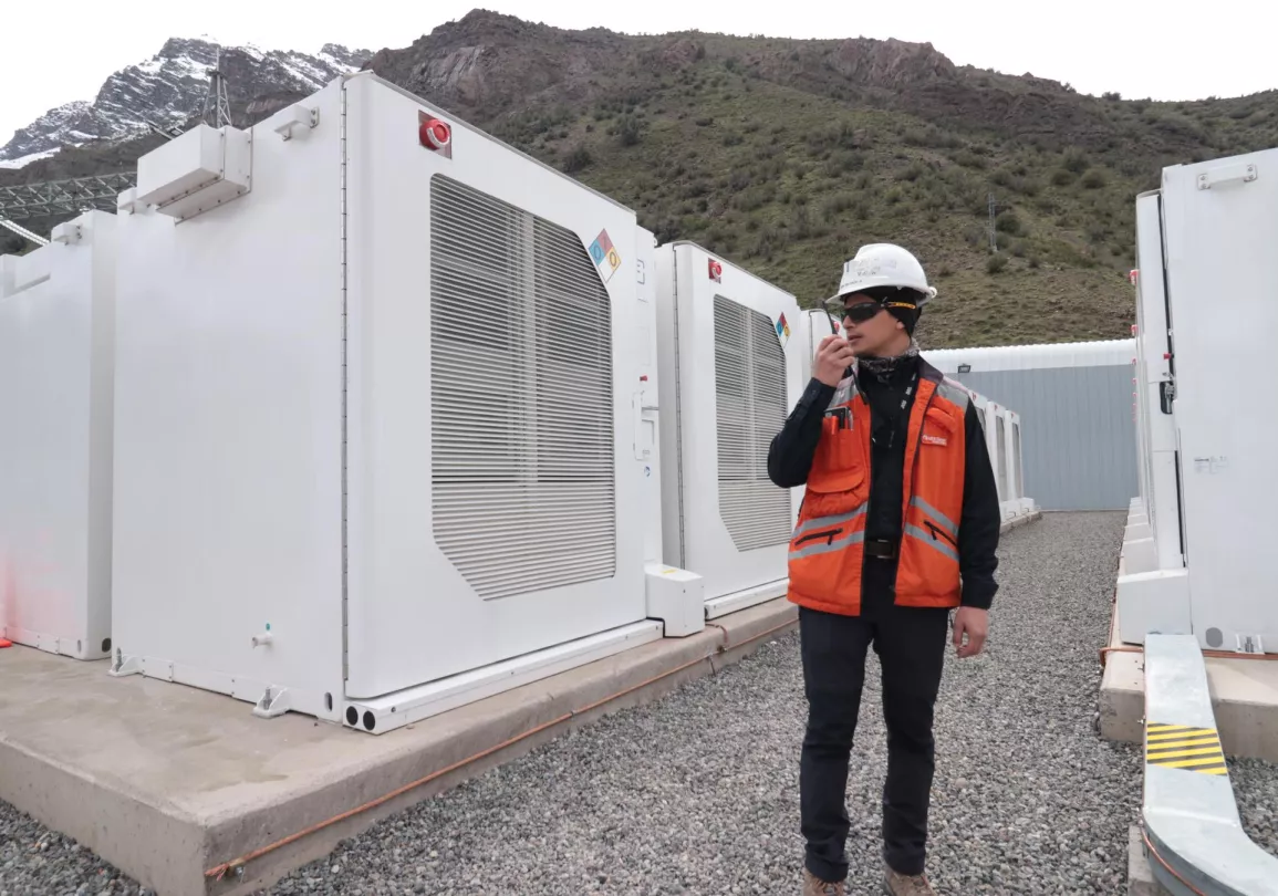 A person wearing a hard hat and orange safety vest holds a radio while standing in front of large white industrial units at a power facility. There are mountains in the background.