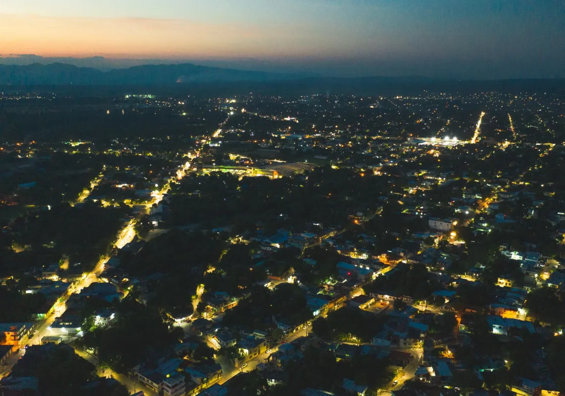 Aerial view of a city at dusk, with streets illuminated by streetlights. The city is densely packed with buildings, and the horizon shows a faint glow from the setting sun, with mountains in the distance.