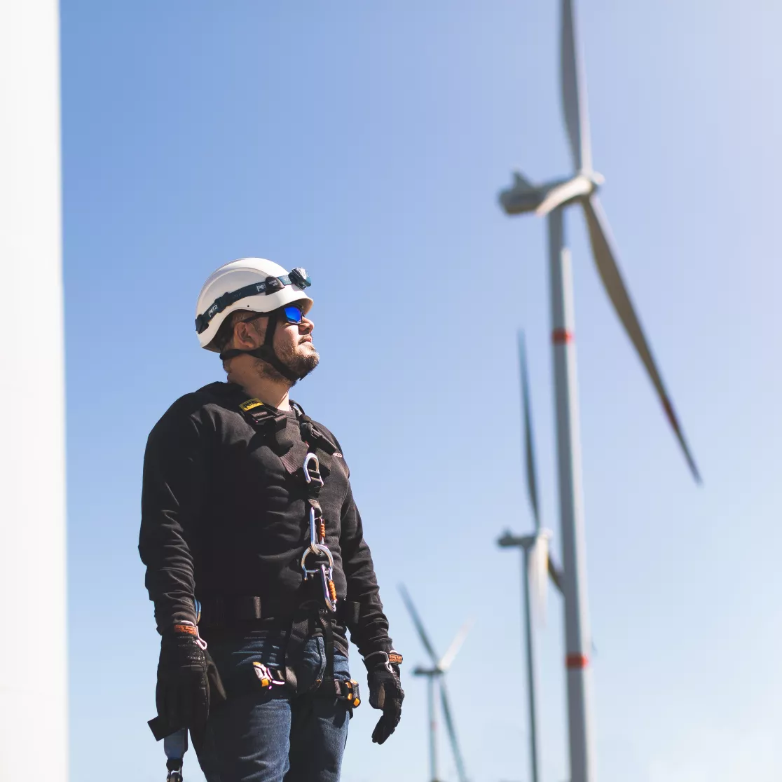 A worker wearing safety gear and a helmet stands near wind turbines under a clear blue sky.