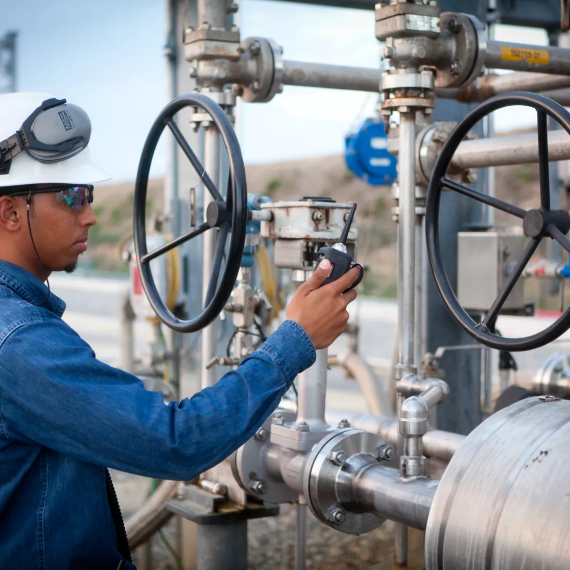 An industrial worker wearing a hard hat and safety glasses operates machinery with large valves outdoors.