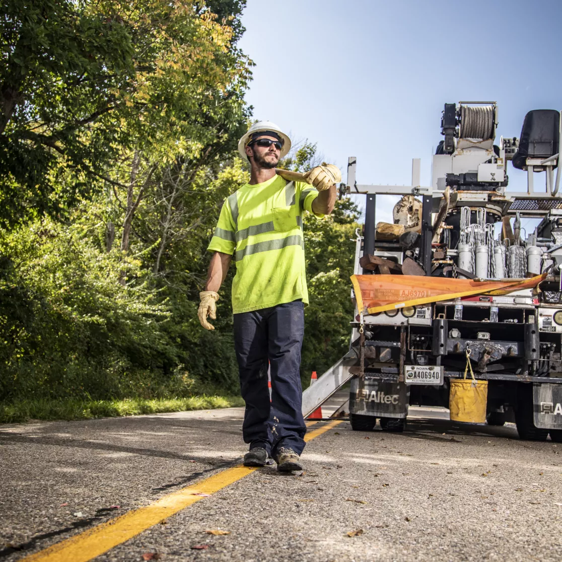 A construction worker in a neon yellow safety vest and helmet walks along a road with a truck in the background. The road is lined with trees under a clear blue sky.
