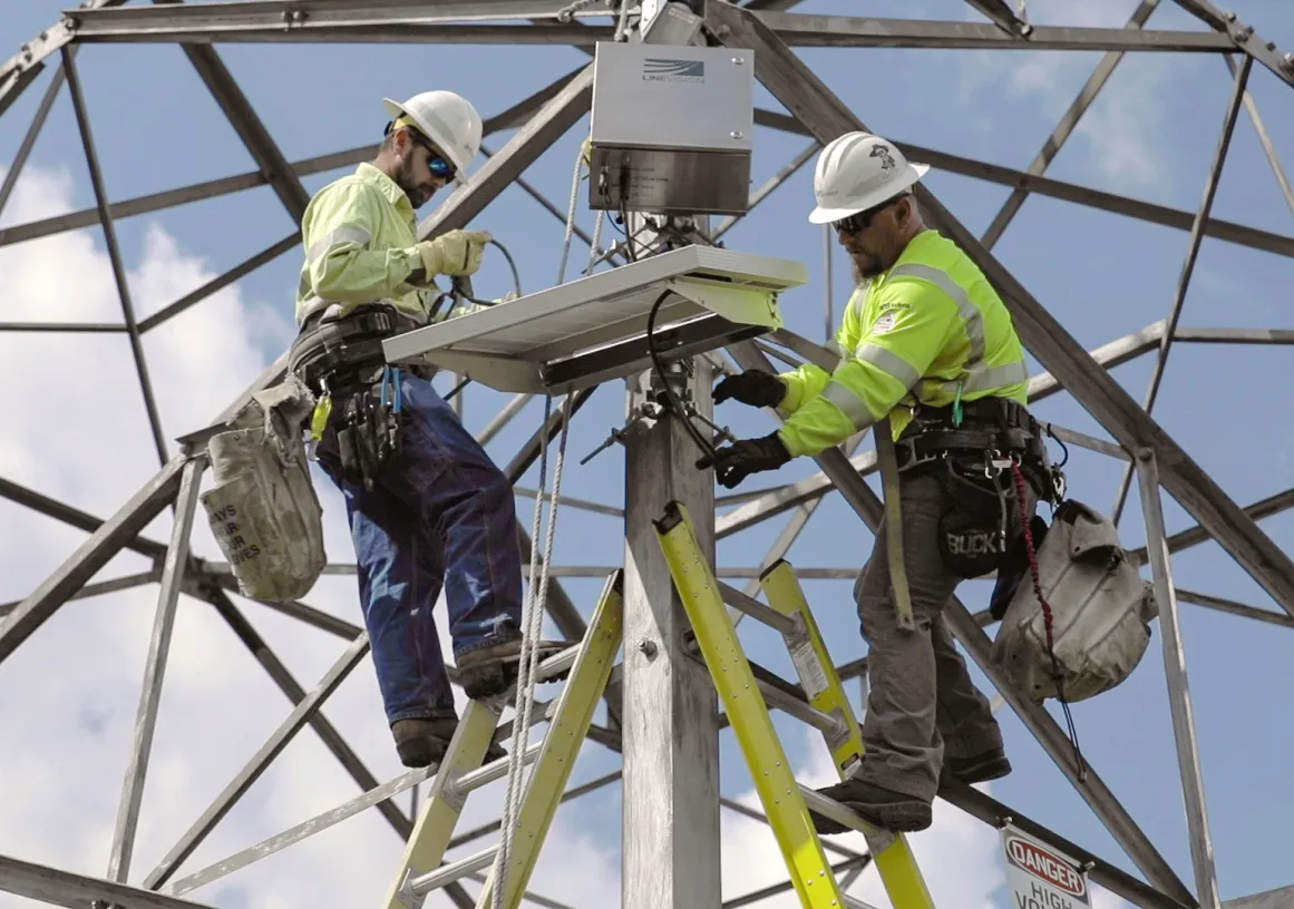Two utility workers in safety gear and helmets are installing equipment on a metal tower. They are using ladders and are surrounded by a framework of steel beams against a blue sky.
