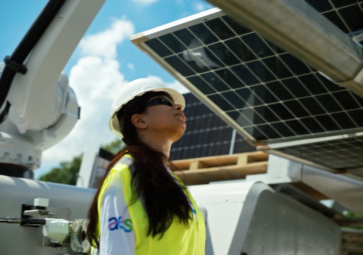 A person wearing a white hard hat, safety glasses, and a yellow safety vest is looking up at a solar panel. The person is standing outdoors with a partly cloudy sky in the background. Industrial equipment is visible around them.