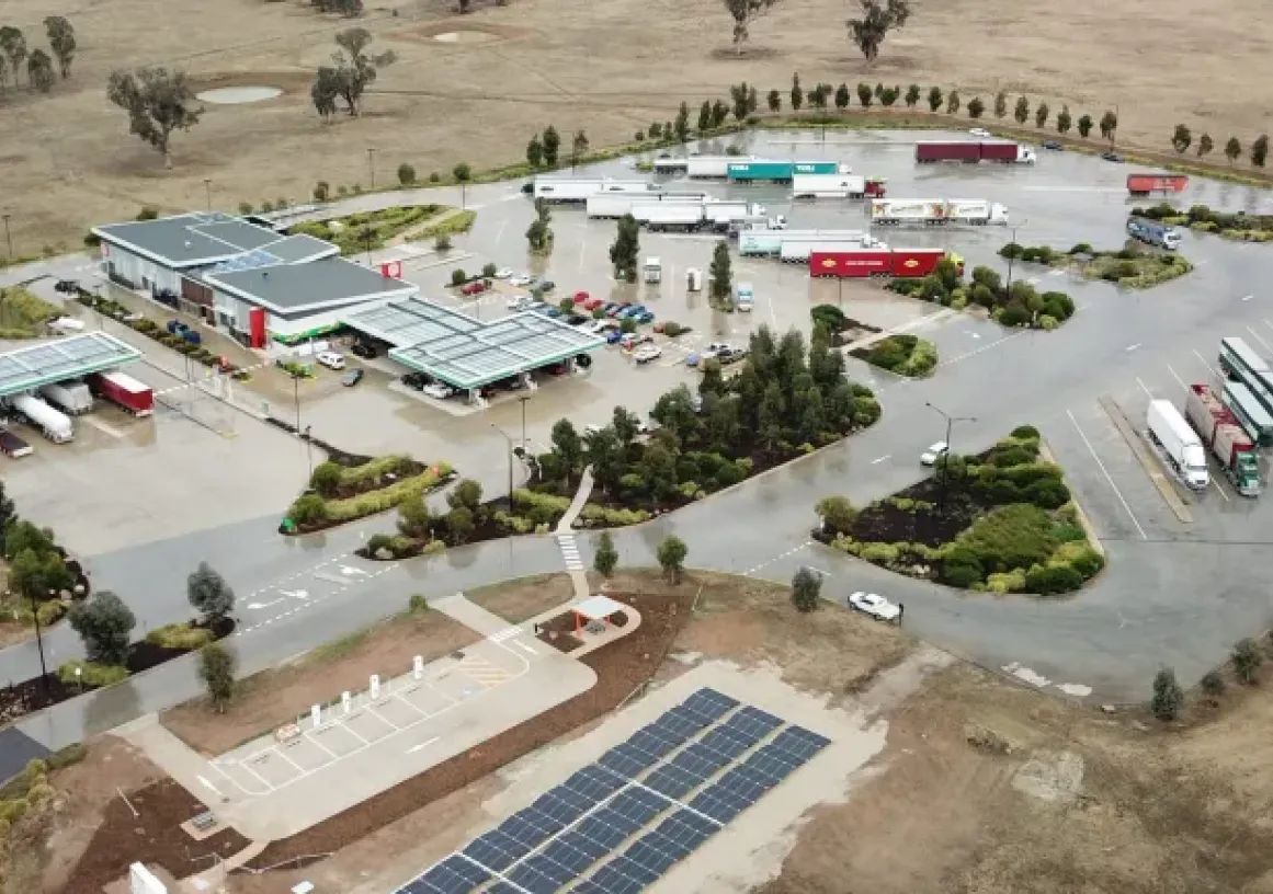 Aerial view of a large truck stop with multiple parked trucks, a convenience store, and fuel pumps. The area is surrounded by open land with scattered trees and features solar panels in the foreground.