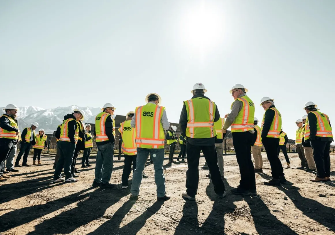 A group of people wearing hard hats and high-visibility vests stand in a circle on a construction site with mountains in the background. The sun shines brightly in the clear sky.