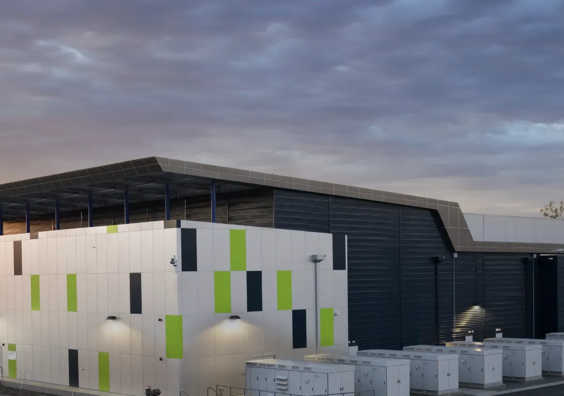 Modern industrial building with a flat roof and colorful rectangular panels on the facade, set against a dramatic cloudy sky at sunset. The structure is surrounded by utility equipment and a fenced perimeter.