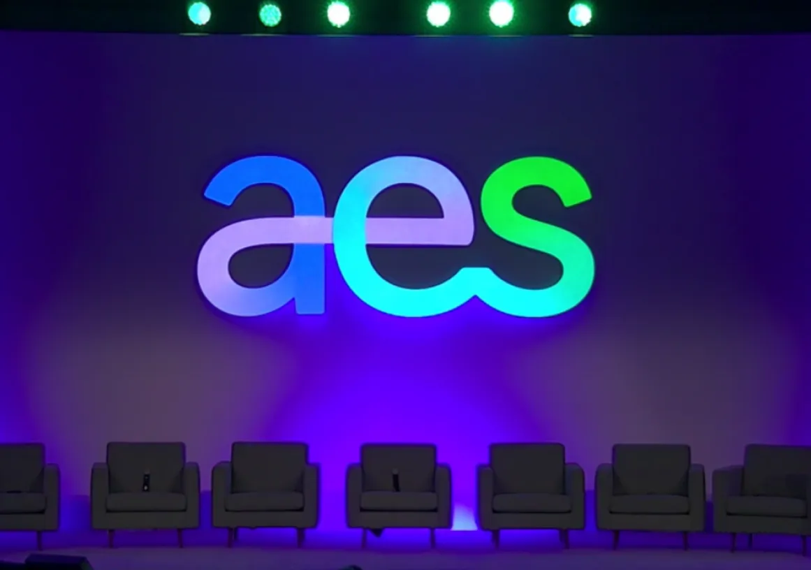 A stage setup with the large letters 'aes' illuminated in blue and green against a purple backdrop. Six empty gray chairs are lined up in front of the backdrop, and a podium with a microphone is on the right side.
