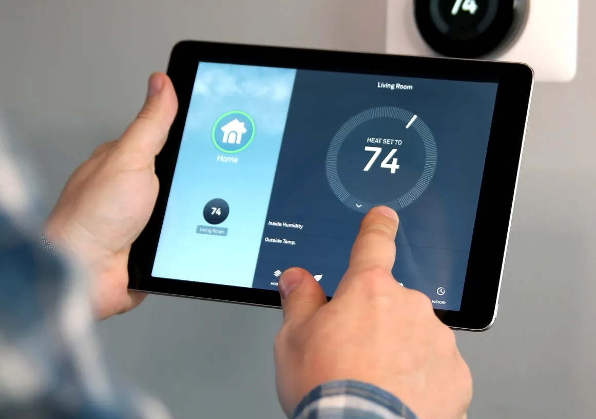 A person adjusts a smart thermostat using a tablet. The tablet screen shows a home automation app with the temperature set to 74 degrees for the living room. A wall-mounted thermostat displays the same temperature.