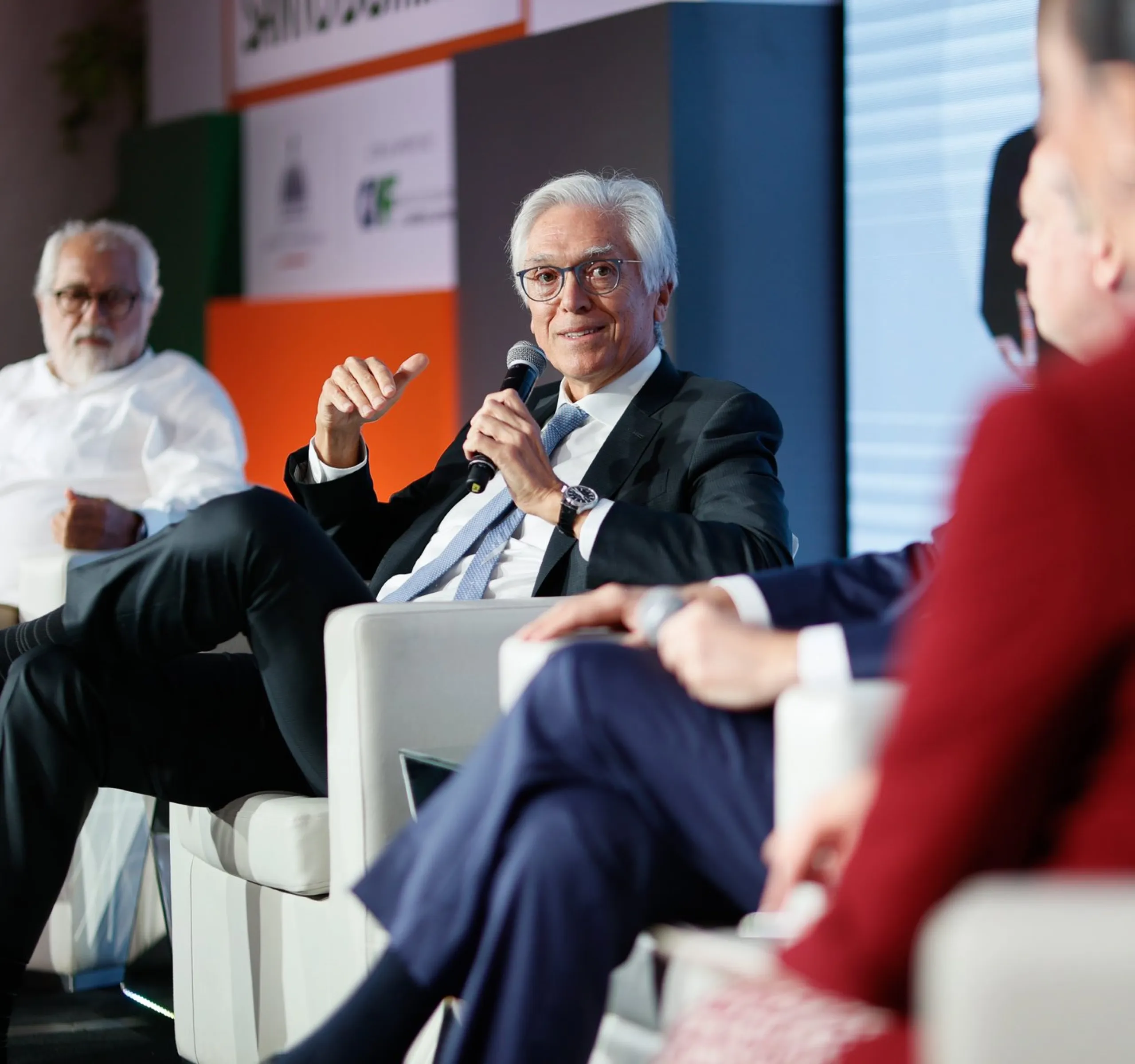 Four people seated on stage during a panel discussion. One man in a suit speaks into a microphone, while others listen attentively. The background displays a colorful banner.