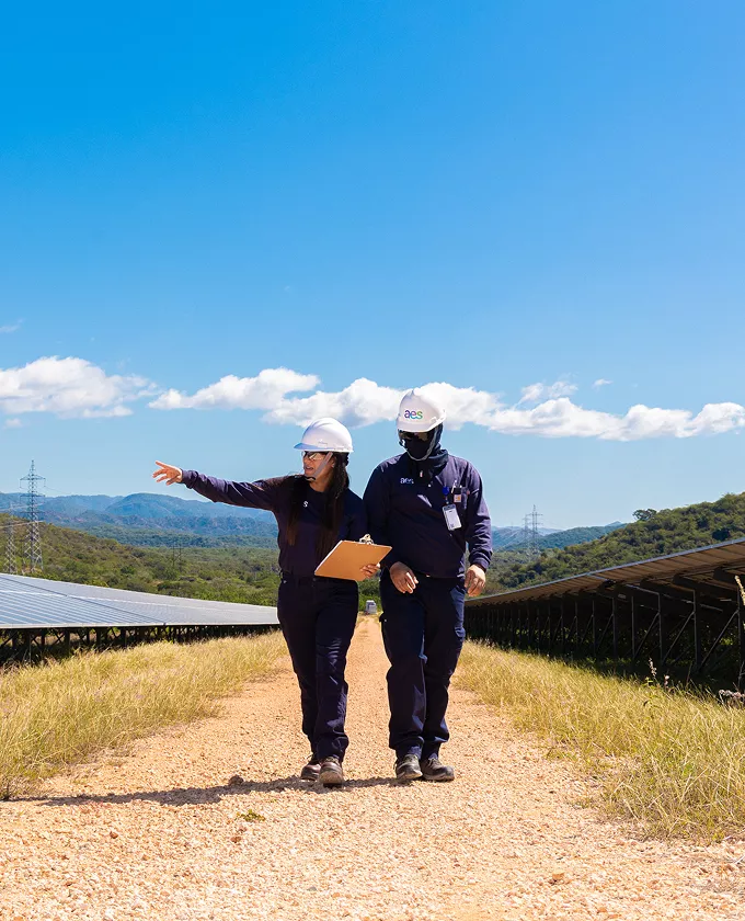Two people wearing hard hats and dark uniforms walk along a gravel path between rows of solar panels. One person is pointing forward, holding a clipboard. The sky is clear with a few clouds, and hills are visible in the background.