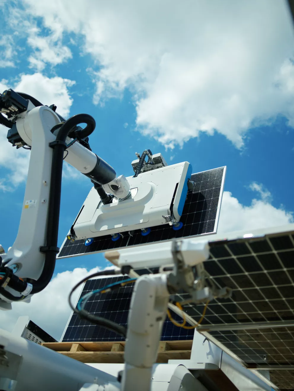 A robotic arm is positioned underneath a solar panel, with a clear blue sky and scattered clouds in the background. The arm appears to be part of a solar panel cleaning system.