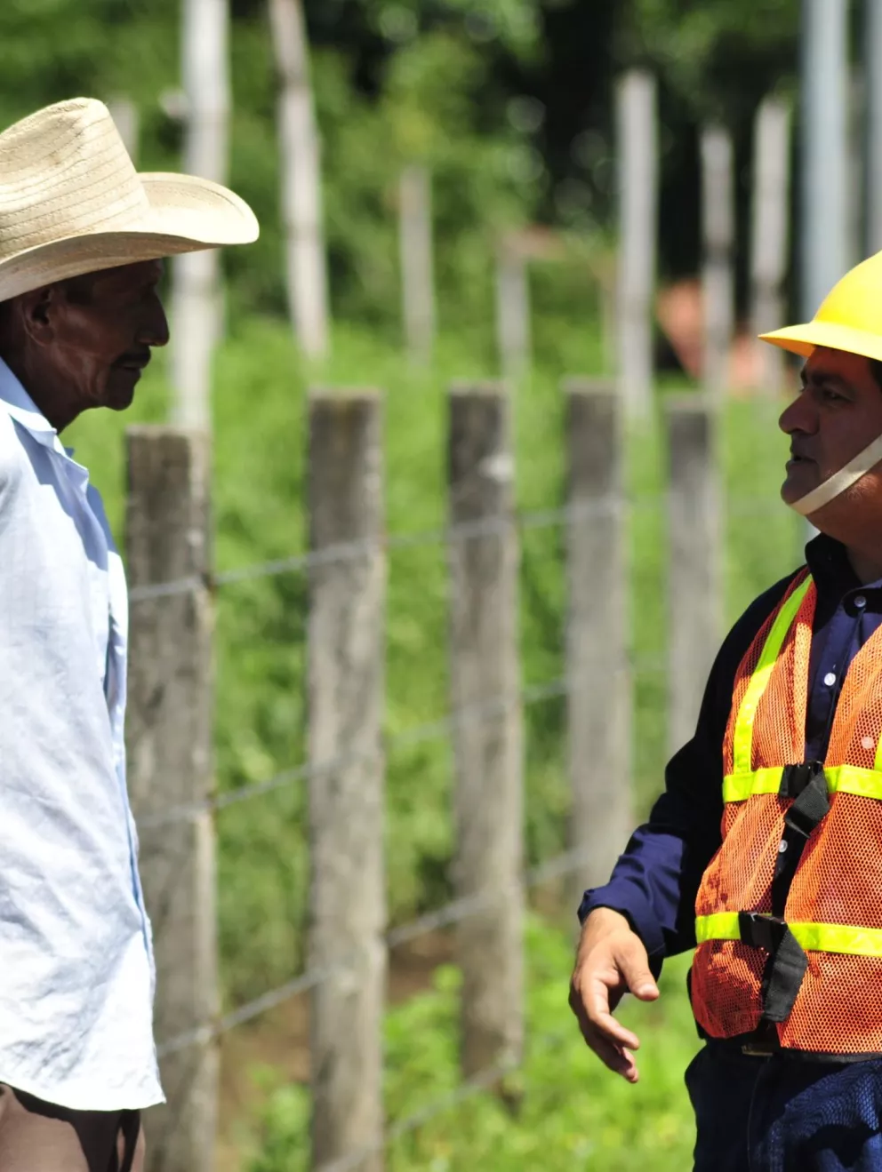 A man wearing a straw hat and light blue shirt stands facing a man in a hard hat and orange safety vest. They are outdoors near a fence with green foliage in the background.