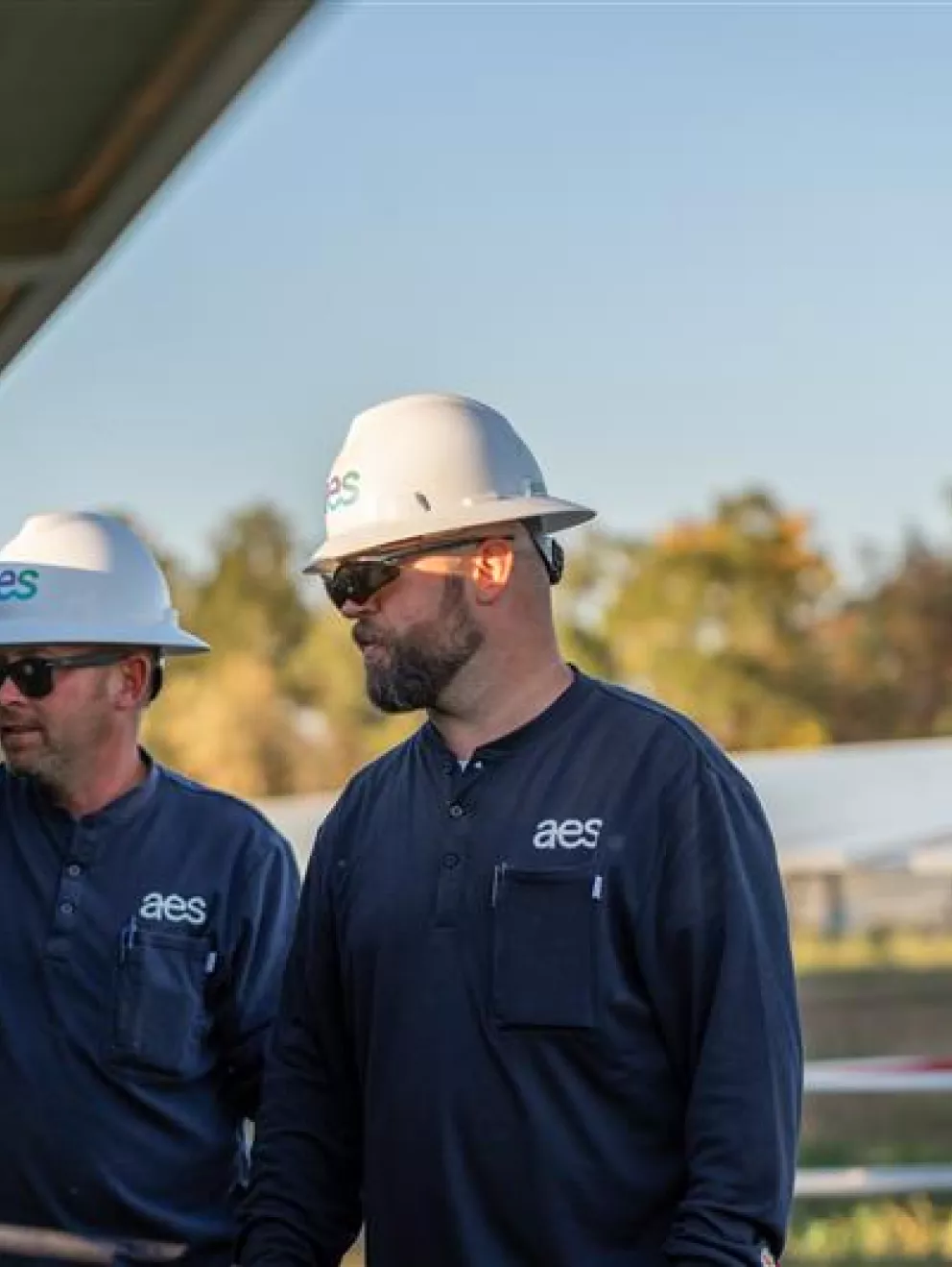 Two workers wearing hard hats and sunglasses stand beside a solar panel installation. They are dressed in navy shirts with 'aes' logos. The background shows more solar panels and trees under a clear sky.