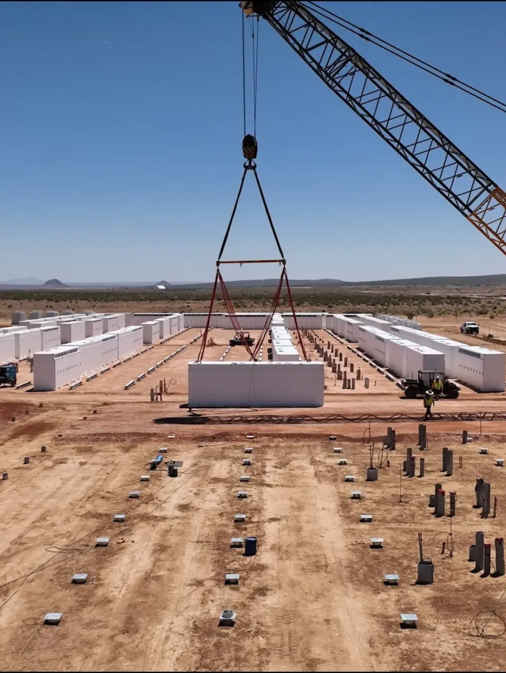 A construction site in a desert landscape features a large crane lifting a white rectangular structure. Several white structures are aligned in rows, with construction vehicles and workers visible on the dirt ground. The sky is clear and blue, with distant hills on the horizon.