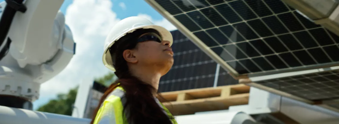 A person wearing a white hard hat, safety glasses, and a yellow safety vest is looking up at a solar panel. The person is standing outdoors with a partly cloudy sky in the background. Industrial equipment is visible around them.
