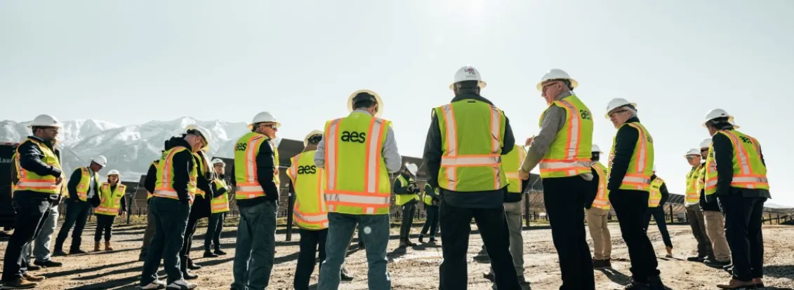 A group of people wearing hard hats and high-visibility vests stand in a circle on a construction site with mountains in the background. The sun shines brightly in the clear sky.