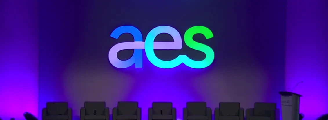 A stage setup with the large letters 'aes' illuminated in blue and green against a purple backdrop. Six empty gray chairs are lined up in front of the backdrop, and a podium with a microphone is on the right side.