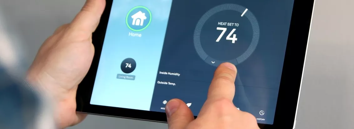 A person adjusts a smart thermostat using a tablet. The tablet screen shows a home automation app with the temperature set to 74 degrees for the living room. A wall-mounted thermostat displays the same temperature.