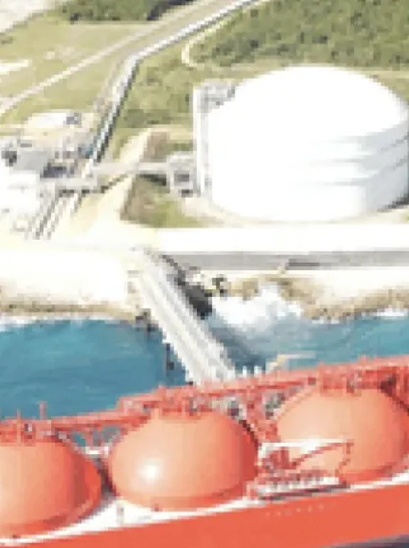 Aerial view of a large red liquefied natural gas (LNG) tanker docked at a coastal facility. The facility includes a large white storage tank and infrastructure surrounded by green vegetation. The sea is a vibrant blue, and the coastline is rocky.