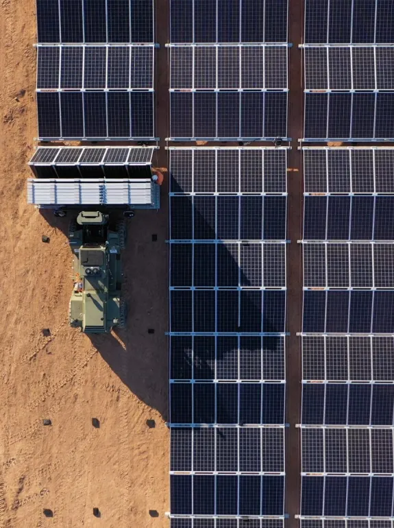 Aerial view of a solar farm with rows of solar panels on the right and a vehicle with equipment on the left in a sandy area.