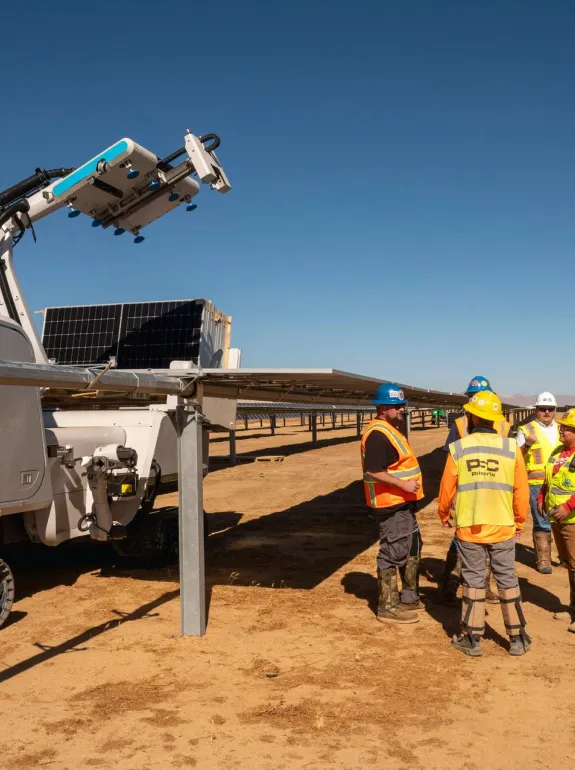 A group of workers in safety vests and helmets stand near a robotic machine labeled 'maximo aes' at a solar farm. The machine is on tracks and positioned next to solar panels under a clear blue sky.