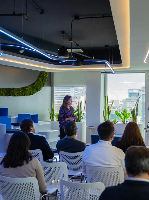 A woman stands in front of an audience in a modern conference room, presenting a slide about the Paris Agreement. The room features large windows with city views, plants, and contemporary lighting. Attendees are seated on white chairs, facing the presenter.