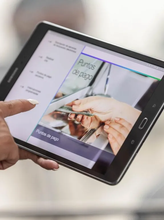 A person holds a tablet displaying a screen with the text 'Puntos de pago' and an image of hands exchanging a credit card. The person is using their finger to interact with the tablet screen.