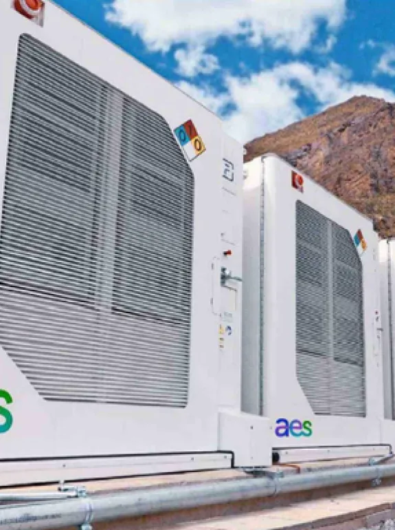 A worker wearing a hard hat and orange safety vest walks beside large white industrial units with the AES logo. The units are set outdoors with mountains and a blue sky in the background.