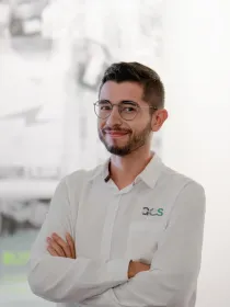 A person with glasses and a beard wearing a white shirt with the AES logo stands with arms crossed, smiling. The background is blurred with some text and images visible.