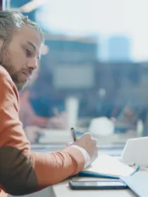 A person with a beard and gray hair is sitting at a desk, wearing an orange sweater and using a pen to write in a notebook. They have an earbud in one ear and are surrounded by a laptop, a smartphone, and a face mask. The background shows a blurred office environment with large windows.