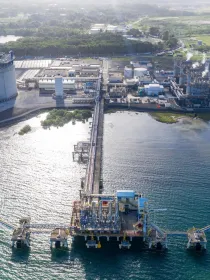 Aerial view of an industrial facility by the water, featuring a large cylindrical tank, multiple buildings, and a long pier extending into the sea. The facility is surrounded by green fields and trees, with smoke visible from some chimneys.