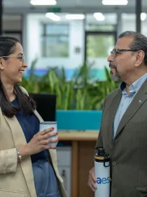 Two colleagues in an office setting, a woman in a beige blazer holding a coffee cup, and a man in a green suit with a water bottle, engaging in conversation.