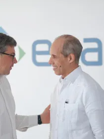 Two men in white shirts are standing and talking in front of a blurred logo background. The man on the left is gesturing with his hand on the other man's shoulder, and both are smiling.