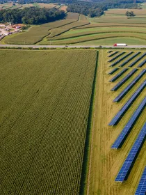 Aerial view of a landscape featuring rows of solar panels on a grassy area adjacent to a large field of crops. A road separates the solar panels from the crops. In the background, there are more fields and trees, with a few buildings visible on the left side.