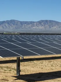 A large array of solar panels installed in a desert landscape with a backdrop of distant mountains under a clear blue sky.