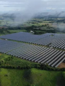 Aerial view of a large solar farm with rows of solar panels surrounded by lush green fields and hills. Misty clouds hover above the landscape, and distant mountains are visible under a cloudy sky.