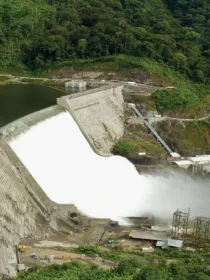 Aerial view of a large dam with water flowing over its spillway, surrounded by lush green forest. The dam is curved and made of concrete. A mist forms at the base where the water impacts. Roads and small buildings are visible near the dam.
