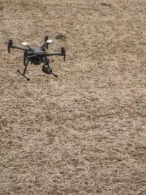 Two people in safety gear operate a drone over a dry field. One person holds a remote control, while the other observes. The drone is flying low above the ground.